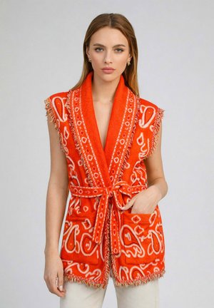 Femme portant un gilet-robe sans manches orange à motifs, avec ceinture nouée à la taille et grandes poches avant, se tenant devant un fond uni.