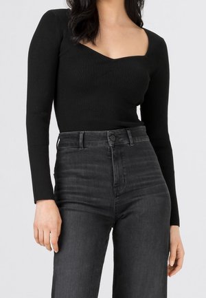 Vrouw draagt een zwart geribbeld shirt met lange mouwen en sweetheart-halslijn, gecombineerd met een hooggetailleerde donkere grijze jeans, staand tegen een effen achtergrond.