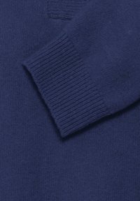 Tumši zilā adīta džemperis ar ribotām aprocēm, parādot mīkstu un ērtu auduma tekstūru.