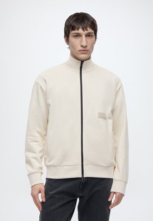 Uomo che indossa una felpa beige con collo alto e zip e pantaloni neri, in piedi davanti a uno sfondo chiaro uniforme.