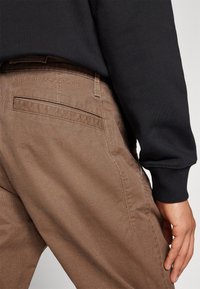 Bruna texturerade byxor med en subtil väv. Har en bakficka och ett präglat logotyp på midjebandet. Kombination med en svart långärmad skjorta.