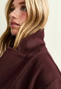 Cappotto in misto lana borgogna con colletto alto, texture liscia, caratterizzato da un design pulito e una silhouette strutturata, mostrato di lato.
