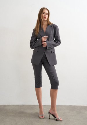 Kvinde iført en mørkegrå oversized blazer og matchende knælange bukser står på en grå gulv mod en hvid væg.