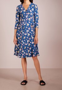 Blå omslagsklänning med ett blommönster i rosa, gult och vitt. Har 3/4-ärmar och en svängd kjol. Matchad med svarta halksandaler.