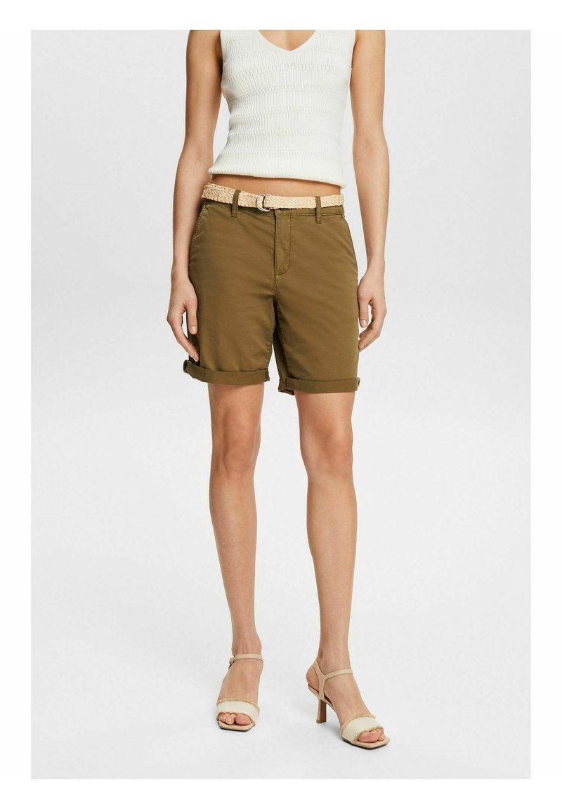 Femme portant un short marron retroussé avec une ceinture beige, un top blanc sans manches et des sandales à talons hauts beiges, debout devant un fond uni.