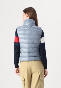 Gilet matelassé bleu clair avec col haut, texture en losanges et sans fermetures visibles, porté sur un pull à rayures bleu marine, rouge et blanc.
