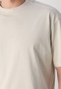 Homme portant un t-shirt uni beige clair à manches courtes avec le texte "BOSS" brodé près de l'encolure sur la poitrine.