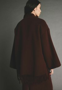 Burgundy gyapjú poncsó laza szabással, széles ujjaival és bojtós szegéllyel. Simafejű textúra, minimális dizájn, és nincs záródás.