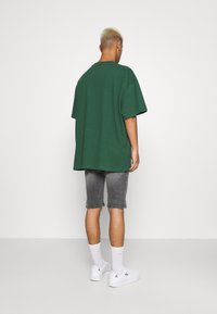 Camiseta verde de gran tamaño, de material de algodón, con mangas cortas, vista trasera. Combinada con pantalones cortos de mezclilla grises y zapatillas blancas. Diseño limpio, sin patrones.