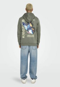 Olijfgroen hoodie met een grote, veelkleurige vlinderafbeelding op de achterkant, gecombineerd met lichtblauwe baggy jeans en witte sneakers.