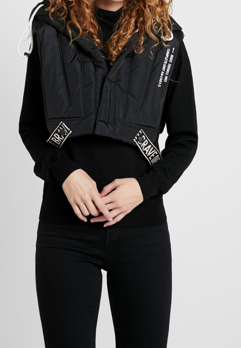 Svart pufferväst med en kortad design, featuring en hög krage, frontdragkedja och sidopatchar med tryckt text. Fästa dragsnören.