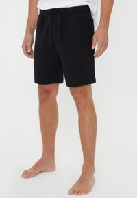 Svarta bomullsshorts med elastisk midja och dragsko. Slät textur, rak skärning och knälängd, bärs barfota.