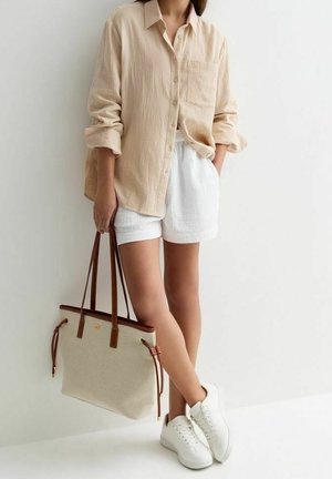 Personne portant une chemise beige à boutons, un short blanc, des baskets blanches, tenant un sac fourre-tout beige avec des anses en cuir marron, debout contre un mur blanc.