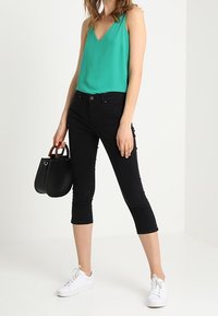 Žena v zeleném tílku, černých krátkých kalhotách, bílých teniskách a držící černou kabelku s hnědými rukojeťmi.