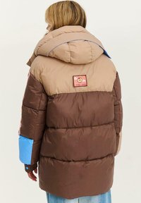 Osoba v farebne rozlíšenom zateplenom kabáte v béžovej, hnedej a modrej farbe s kapucňou a nášivkou "Sweet Things" na chrbte.