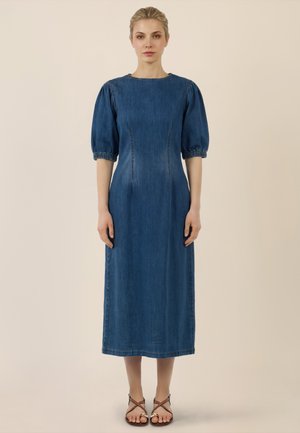 Kobieta o blond włosach nosi niebieską sukienkę midi z denimu z bufkami i rękawami sięgającymi do łokcia oraz brązowe sandały na paski, stojąc na beżowym tle.