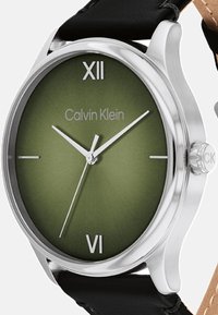 Zegarek Calvin Klein z okrągłą srebrną kopertą, zieloną gradientową tarczą, czarnym skórzanym paskiem oraz znacznikami w postaci rzymskich cyfr przy godzinach 6 i 12.