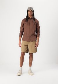 Smeđa košulja dugih rukava s gumbima, bež šorts s teksturom, siva kapa, bijele čarape, bež tenisice. Jednostavan, ležeran outfit.