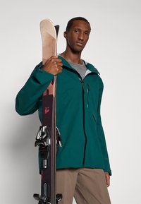 Muškarac u zelenoj jakni drži par skija uspravno, nosi bež hlače, nasuprot jednostavnoj svijetloj pozadini.