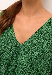 Top de estampado floral verde con escote en V. Tejido ligero que presenta pequeñas flores negras sobre un vibrante fondo verde.