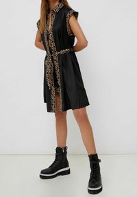 Schwarzes ärmelloses Kleid mit einem gemusterten Kragen und Gürtel. Der Stoff hat eine glatte Textur. Kombiniert mit klobigen schwarzen Schnürstiefeln.