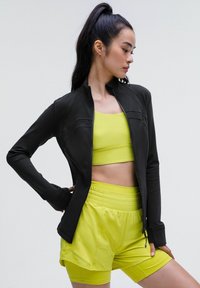 Veste de sport zippée noire au design ajusté, portée par-dessus un crop top jaune vif et un short assorti, le tout en tissu lisse et extensible.