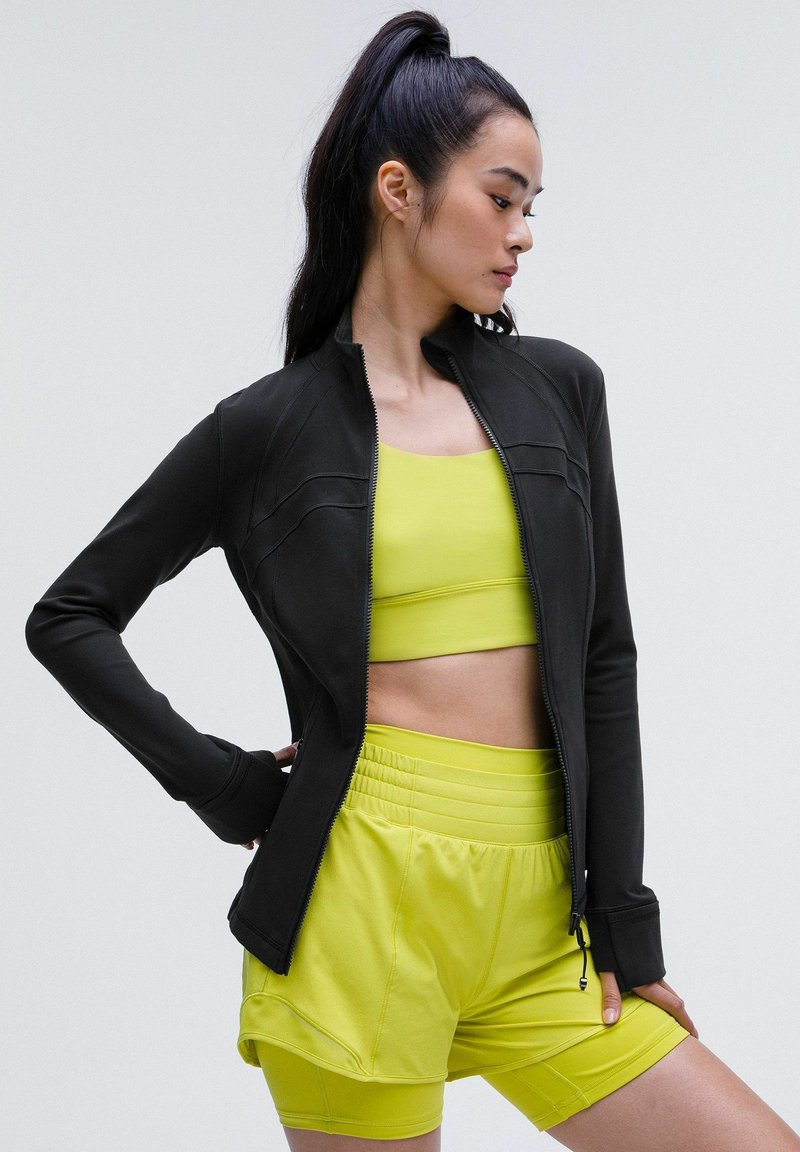 Veste de sport zippée noire au design ajusté, portée par-dessus un crop top jaune vif et un short assorti, le tout en tissu lisse et extensible.