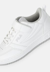 Valkoinen tennari, jossa on tekstuurilla varustettu nahkavarsi, nauha- ja tarranauhakiinnitys, pehmustettu harmaa sisus sekä kumipohja, jossa on pito kuvio.