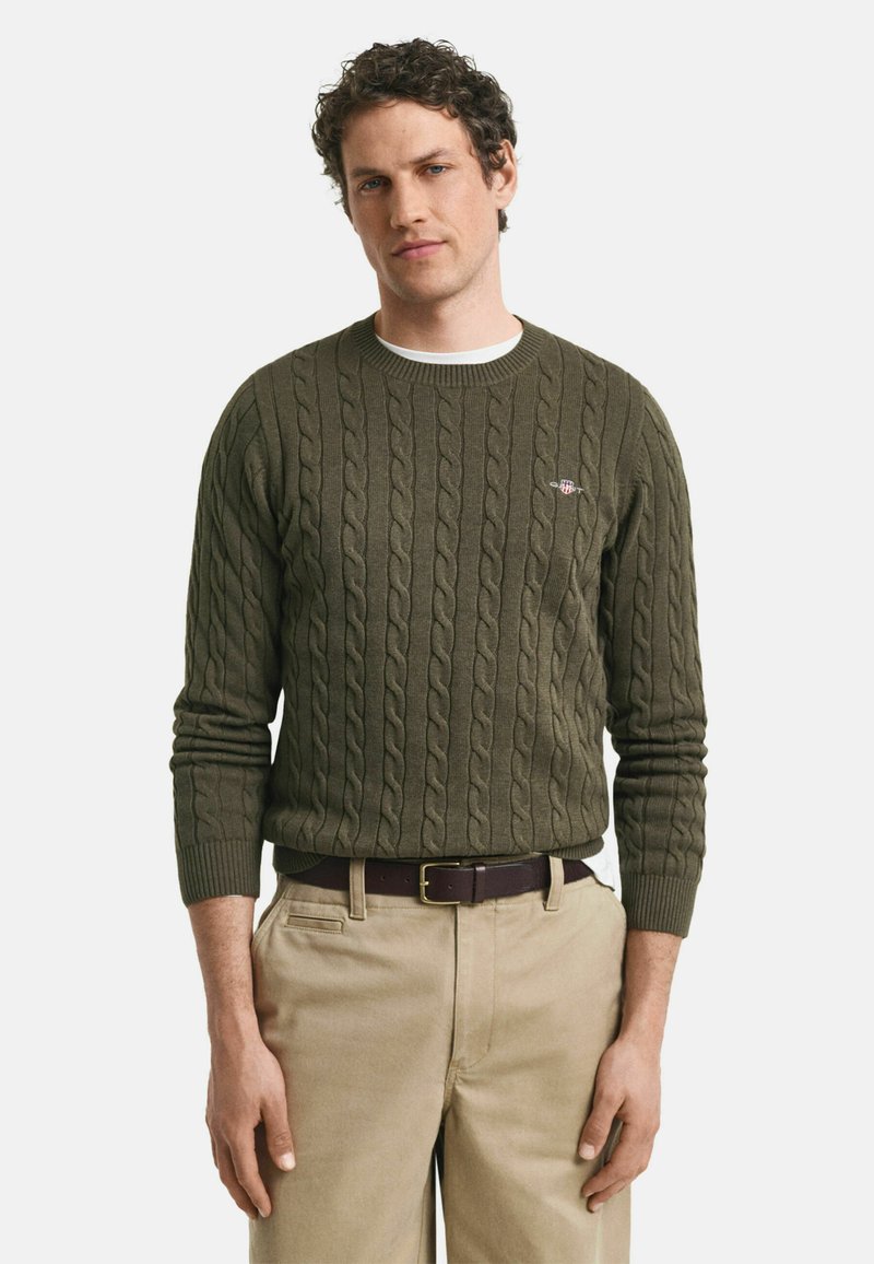 Maglione verde scuro a punto guernsey con collo rotondo e polsini a coste, con un piccolo logo sul petto, abbinato a pantaloni beige.