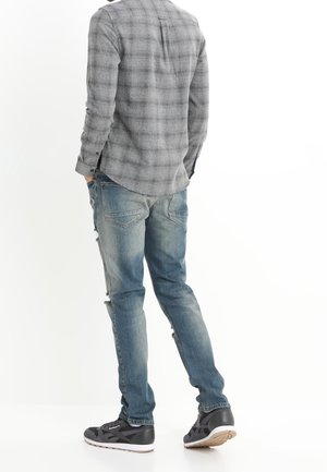Man draagt versleten blauwe spijkerbroek, grijs geruit overhemd en zwarte sneakers, staat met handen in zakken, van achteren gezien, op een witte achtergrond.