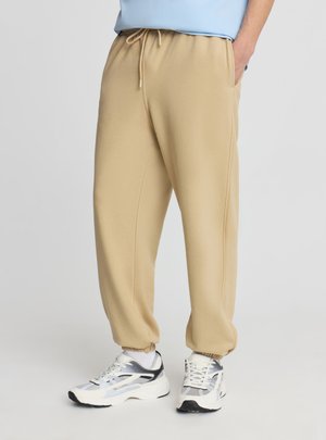 Persona che indossa pantaloni della tuta beige con coulisse e scarpe da ginnastica bianche e grigie, in piedi con una mano in tasca contro uno sfondo semplice.