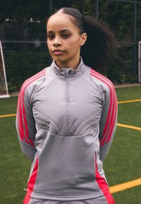Giacca sportiva grigia con colletto alto, caratterizzata da strisce rosa sulle spalle e ai lati. I pannelli testurizzati aggiungono profondità al design.