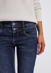 Un jean en denim bleu foncé présente une taille haute avec deux boutons, deux poches avant et une couture contrastante. Un pull léger est porté au-dessus.