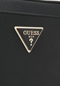 Czarne strukturalne sztuczne skórki z złotym i czarnym trójkątnym logo z napisem "GUESS USA?" oraz znakiem zapytania na środku.