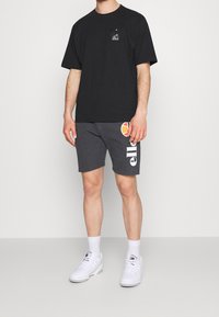 T-shirt en coton noir avec poche et logo, associé à un short gris portant un texte blanc et un design orange. Porté avec des baskets blanches.