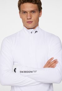 Vit långärmad dragkedjetröja med hög krage, med svarta loggopplikationer på bröstet och ärmen, tillverkad av ett mjukt, stretchigt material.