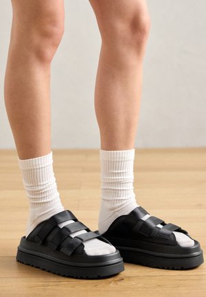 Black platform sandals with three wide straps, textured soles, and open toes, paired with white ribbed crew socks on legs against a light wood floor.
