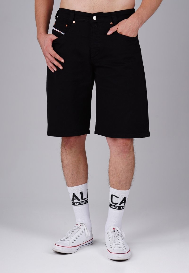 Black cotton shorts with a straight cut, featuring side pockets, a zip fly, and copper hardware accents. Worn with white sneakers and socks.