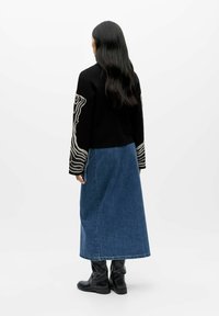 Svart genser med hvit abstrakt broderi på ermene, kombinert med et langt denimskjørt og svarte knelange støvler, sett bakfra.