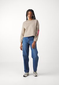 Beige cropped sweatshirt with long sleeves, ribbed cuffs, and a red tag. Paired with blue straight-leg jeans and white sneakers with green accents.
