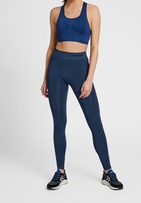 Modrý sportovní top a legíny s vysokým pasem vyrobené z elastického materiálu. Legíny mají texturovaný vzor na stehnech. Černé sportovní boty.