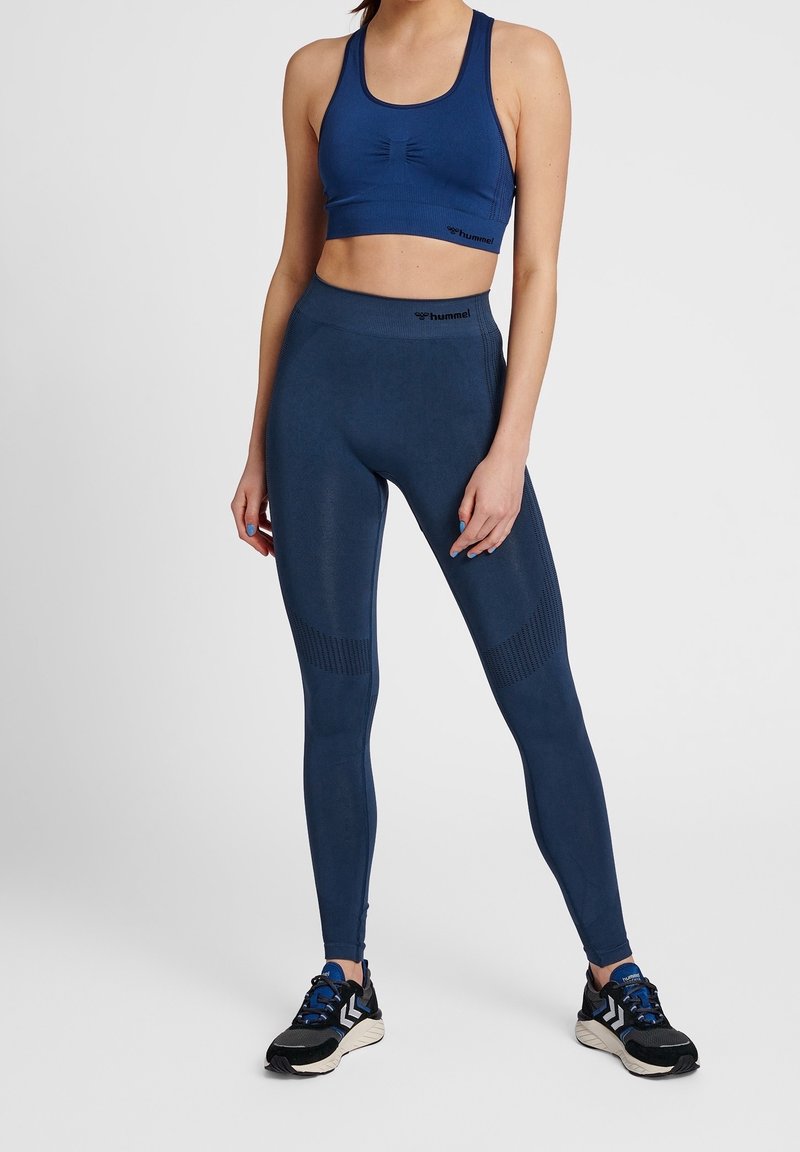 Modrý sportovní top a legíny s vysokým pasem vyrobené z elastického materiálu. Legíny mají texturovaný vzor na stehnech. Černé sportovní boty.