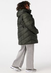 Olijfgroene pufferjack met een capuchon, voorzien van een gewatteerd ontwerp en ritssluiting. Gecombineerd met grijze wijde spijkerbroeken en sneakers.