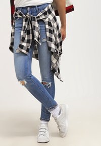 Los jeans skinny rasgados de color azul tienen una cintura alta, combinados con una camisa a cuadros en blanco y negro atada a la cintura. Zapatillas blancas sin cordones.