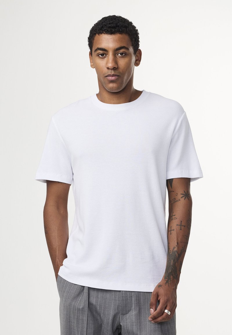 Hvid kortærmet t-shirt med rund hals, lavet af struktureret stof, båret med grå stribede bukser, med synlige tatoveringer på armen.