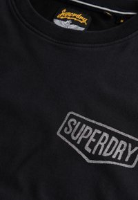 Camiseta negra de algodón. Presenta un logo gris de "SUPERDRY" en la parte frontal, con cuello redondo y detalles cosidos. Etiqueta visible en el interior.