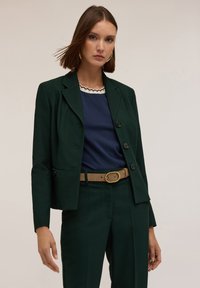 Mörkgrön skräddarsydd blazer med en figursydd design, matchad med en marinblå topp med en vågig halsringning och beige bälte. Mjuk tygstruktur.