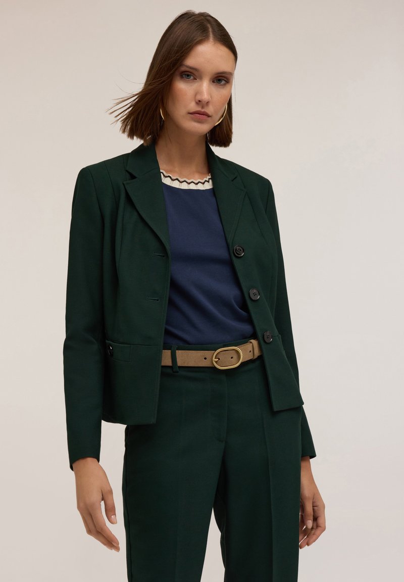 Mörkgrön skräddarsydd blazer med en figursydd design, matchad med en marinblå topp med en vågig halsringning och beige bälte. Mjuk tygstruktur.