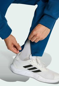 Blaue Sporthose wird über einem weißen Sneaker angepasst. Der Sneaker hat schwarze Streifen und wird mit einer weißen Socke kombiniert.