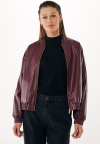 Giacca bomber in finta pelle bordeaux con zip dorata, polsini a costine e vestibilità comoda, indossata sopra un dolcevita nero e jeans neri.
